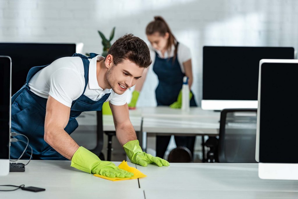 two-young-cleaners-wiping-desks-with-rags-in-offic-NWXGTZE.jpg